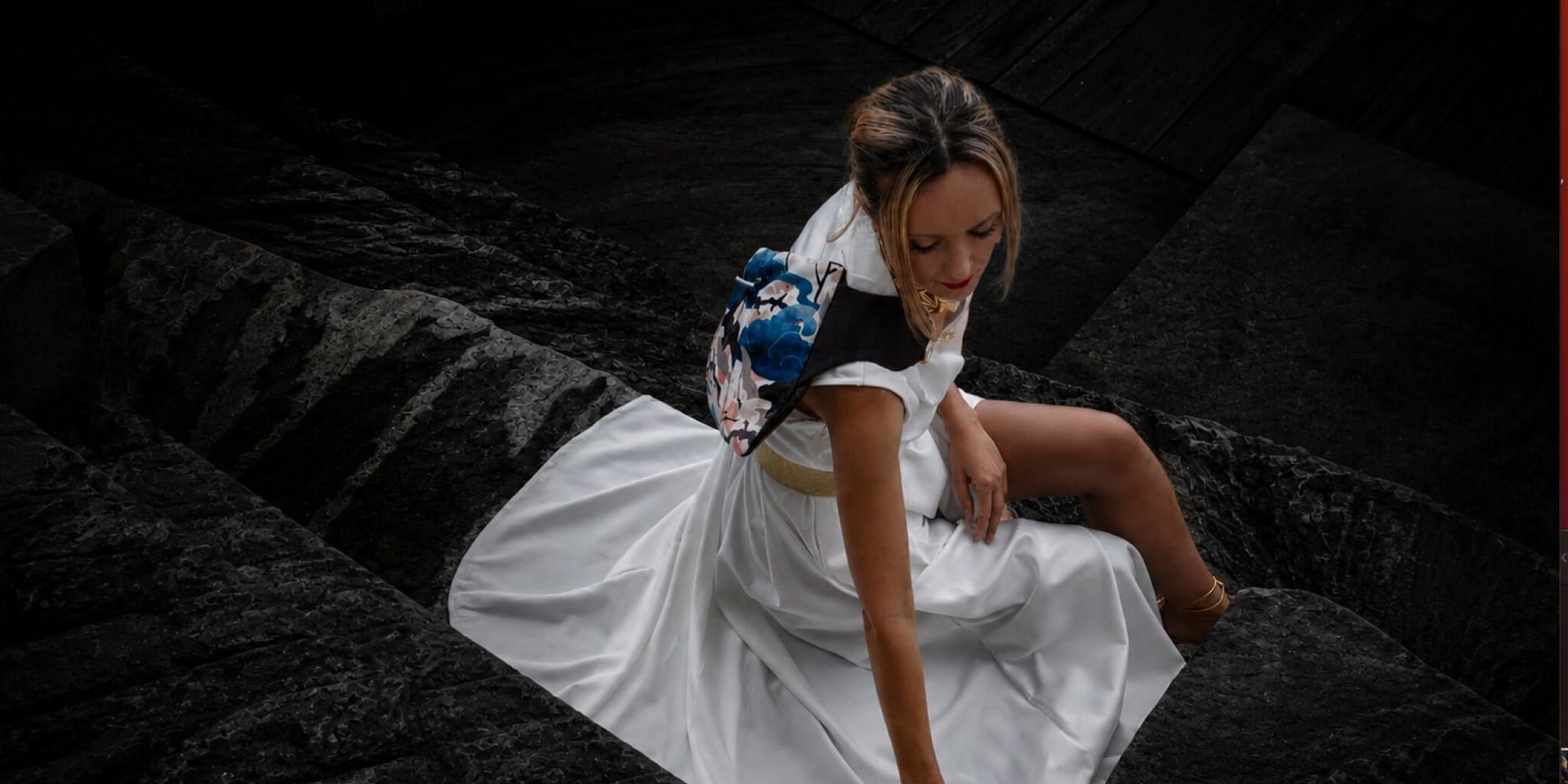 Woman in white dress carrying patterned lvb_collection bag while seated on dark stone steps.