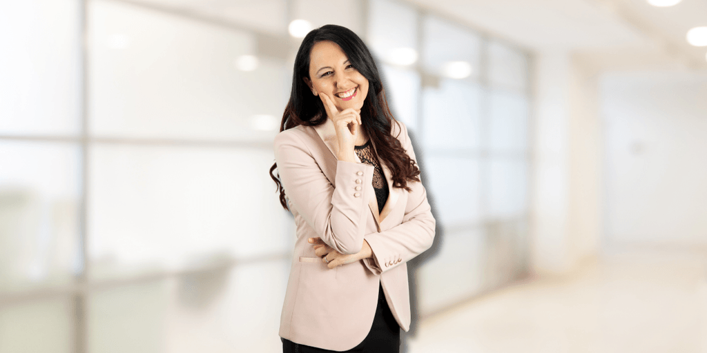 Samya Ilaria Di Donato, a confident woman in a pink blazer, smiles thoughtfully with one hand on her chin.