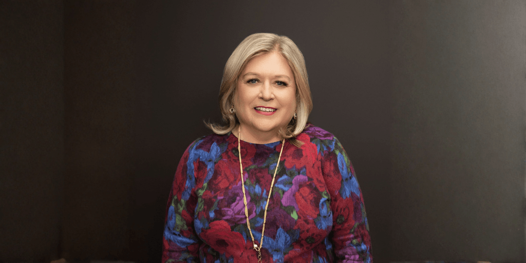 Marcy Westcott, meditation teacher and author, smiling in a colorful floral sweater against a dark background.