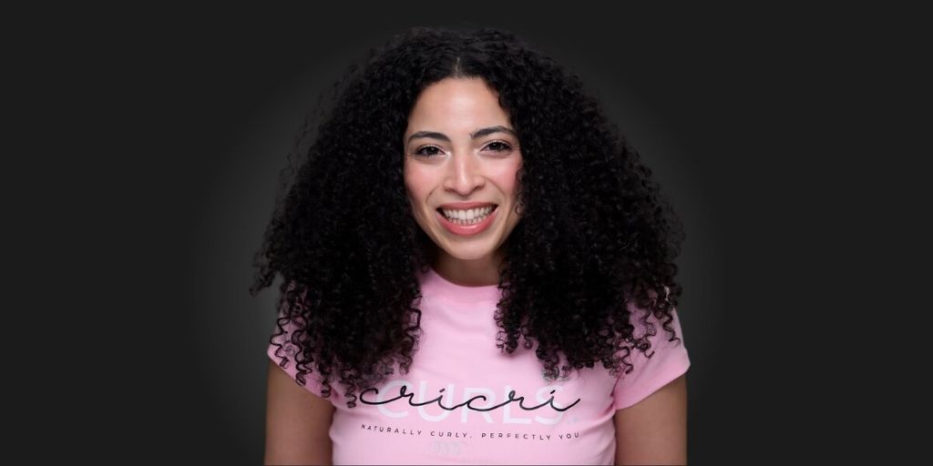 Smiling woman with voluminous curly hair wearing a pink Cricri Curls shirt against a dark background.