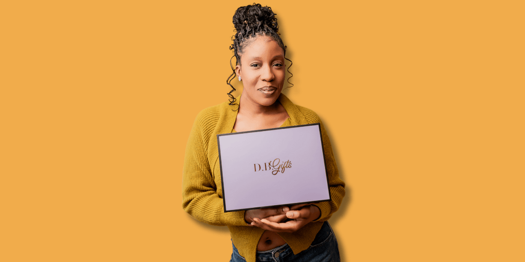 "Smiling woman wearing a yellow sweater, holding a lavender gift box with the D.B. Gifts logo, set against a bright orange background."