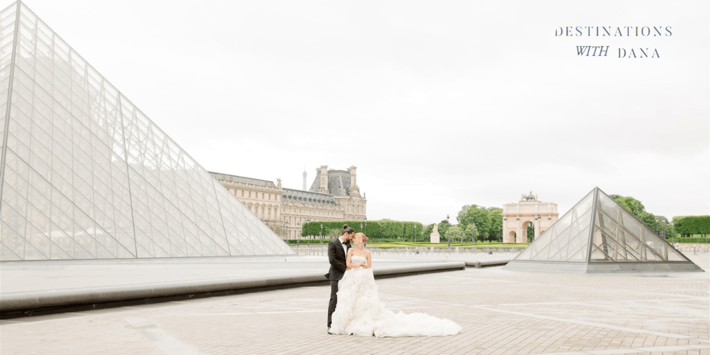 A couple in wedding attire embraces in front of the Louvre Pyramid in Paris, with the Eiffel Tower in the background.