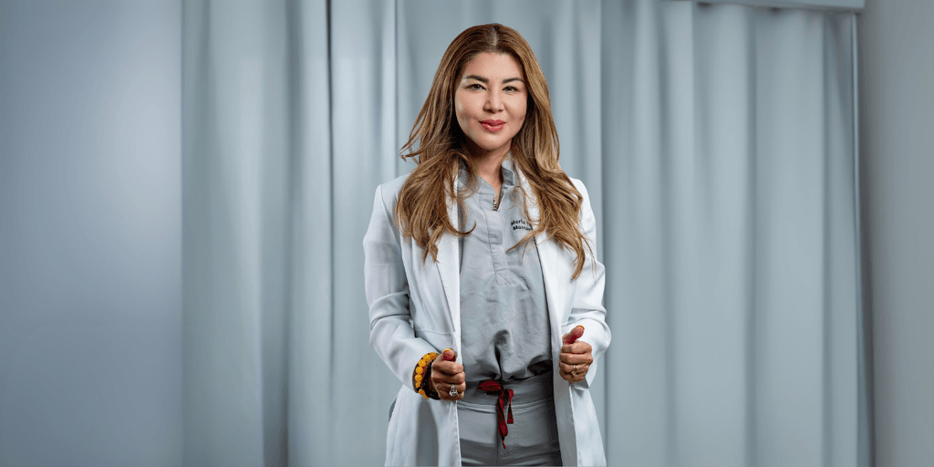 Professional woman in a white lab coat and grey medical scrubs standing confidently with a friendly expression. She is a skilled aesthetician, offering expertise in skincare treatments, with soft lighting and a neutral background.