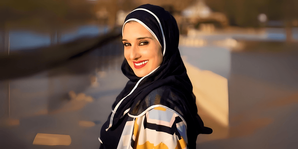 "Smiling woman in a colorful hijab and patterned coat, standing outdoors with a blurred background and sunny lighting."