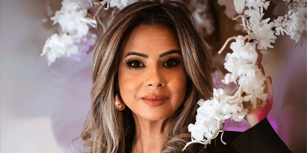 Close-up portrait of a confident woman with glamorous makeup, styled hair, and a warm smile, standing amidst a backdrop of delicate white flowers, radiating elegance and beauty.