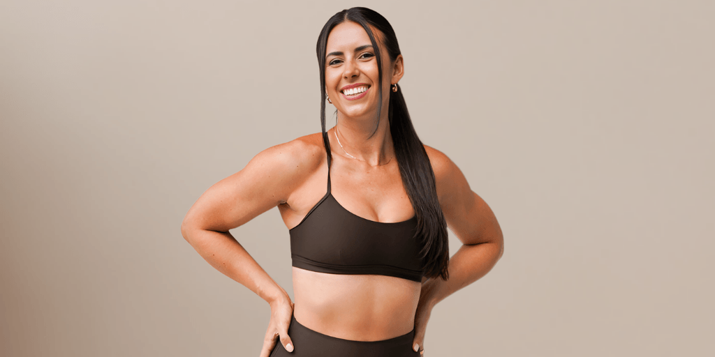 Smiling woman in a dark brown workout outfit, standing with her hands on her hips against a neutral beige background.
