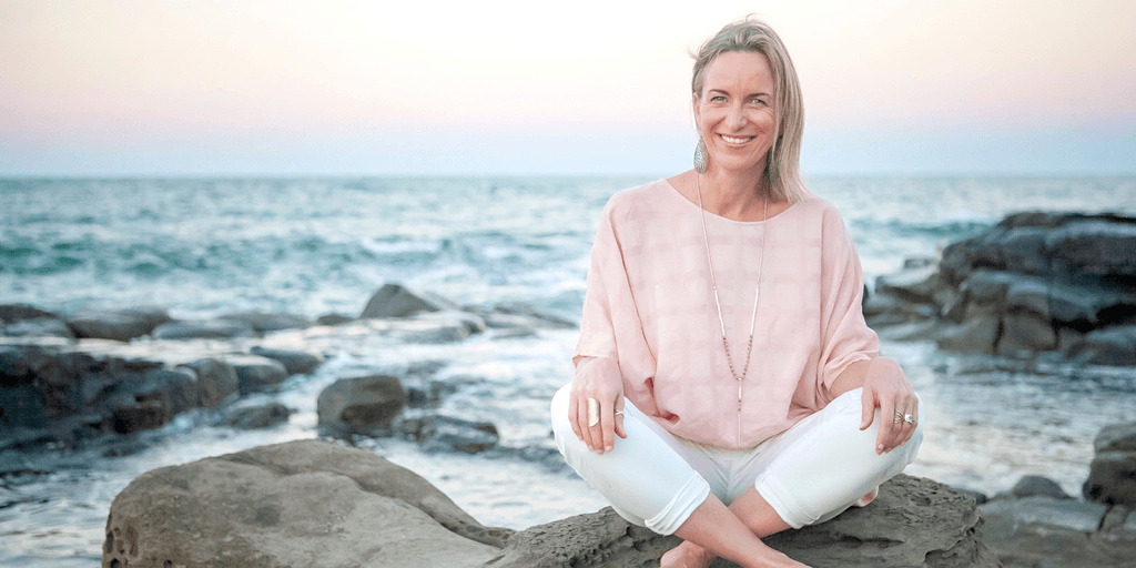 ChatGPT said: Woman in light pink blouse and white pants sitting cross-legged on rocks by the ocean at sunset.