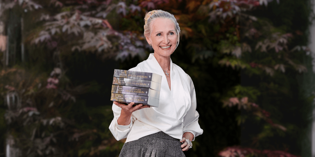 Eva Fellner smiling while holding a stack of The Highlanderess Saga books against a leafy background.