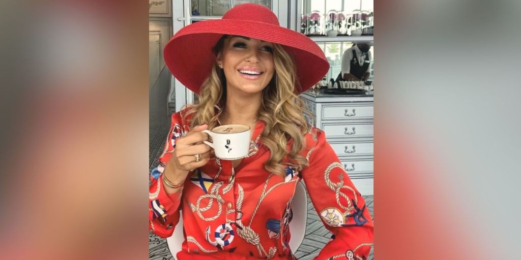 Smiling woman wearing a red hat and a red blouse with a nautical print, holding a coffee cup. She is seated in front of a stylish vanity, creating a chic and fashionable atmosphere."
