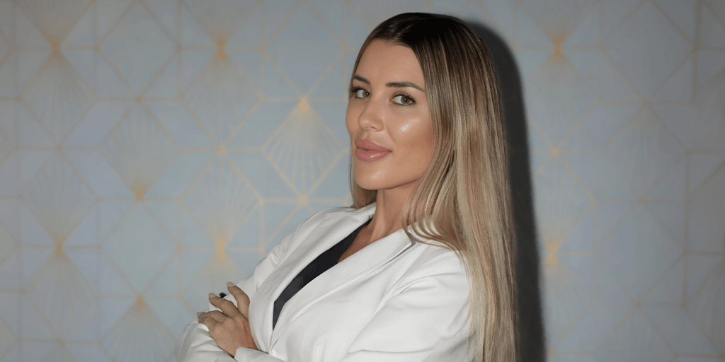 Rita Madozino, founder of Forever Young by Rita, poses confidently in a white coat against a patterned background.