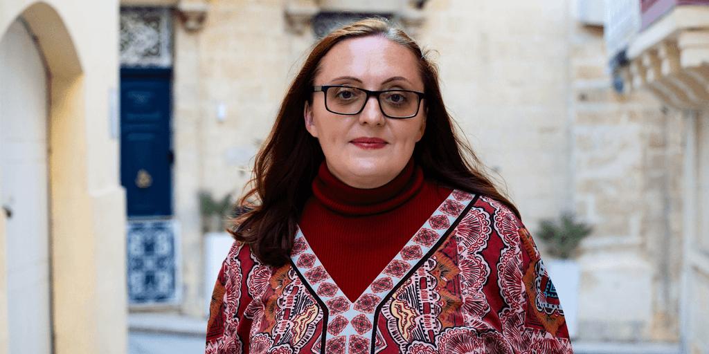 Woman dressed in a red turtleneck and patterned blouse, walking through an old city street with a traditional architectural backdrop.