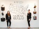 Two women stand in front of a wall displaying bags and a map of the USA
