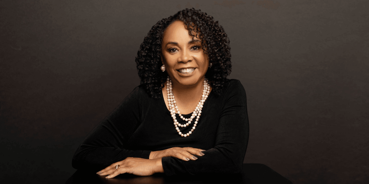 A woman with curly hair, wearing pearls, smiles confidently at the camera.