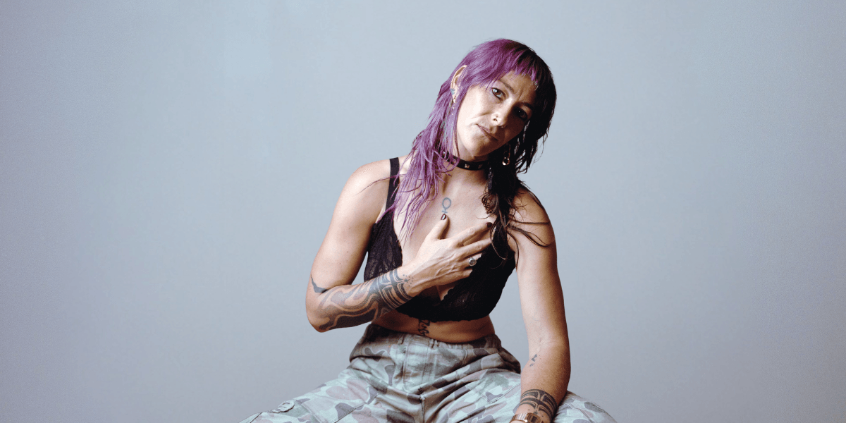 A woman with purple hair and tattoos poses seated against a gray background