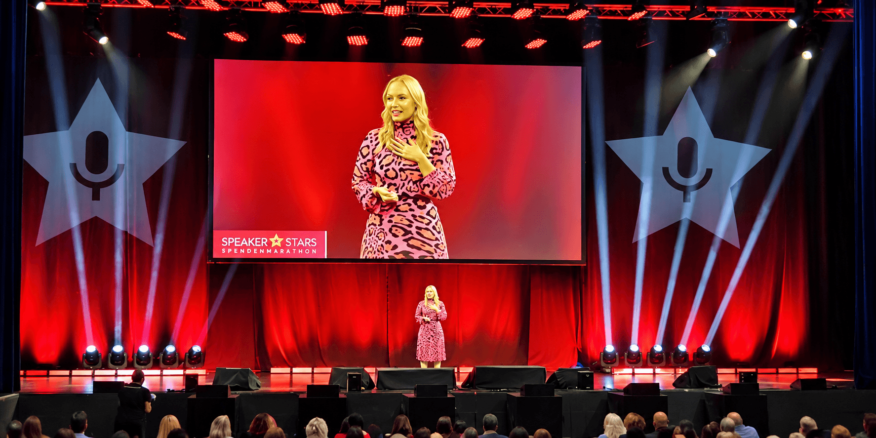 A speaker in a pink dress presents on stage at a conference with an audience.