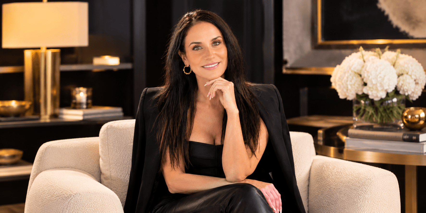 A woman with long dark hair smiles while seated on a couch in a stylish room