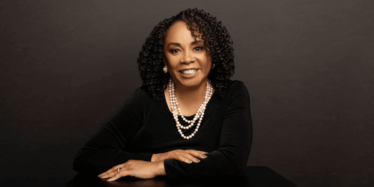 A woman with curly hair, wearing pearls, smiles confidently at the camera.
