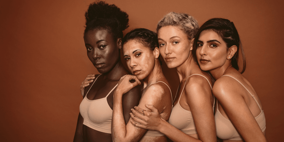 Four women of diverse backgrounds pose together, wearing matching beige tops against a warm backdrop