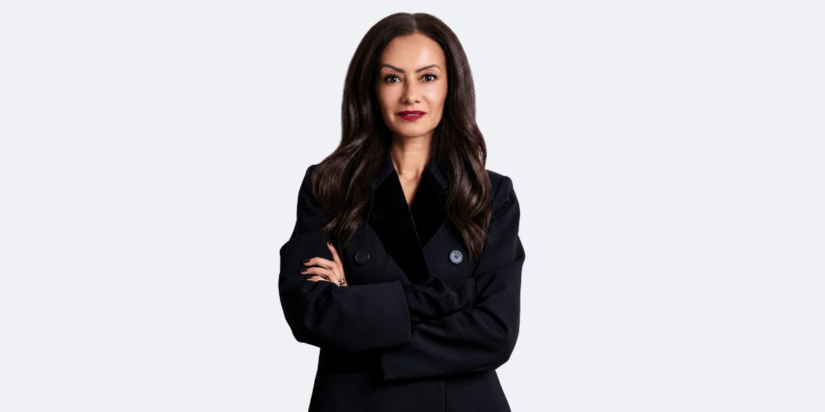 Woman with long dark hair wearing a black coat, arms crossed confidently