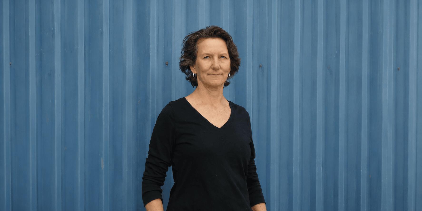 Woman with short hair stands in front of a blue corrugated wall.