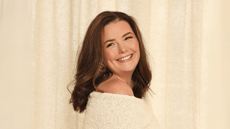 "Portrait of a woman smiling over her shoulder in a soft off-white sweater with wavy brown hair."