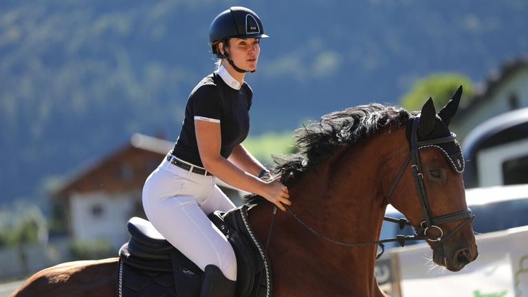 A woman in riding attire competes in showjumping, confidently riding her horse in an outdoor arena surrounded by nature.