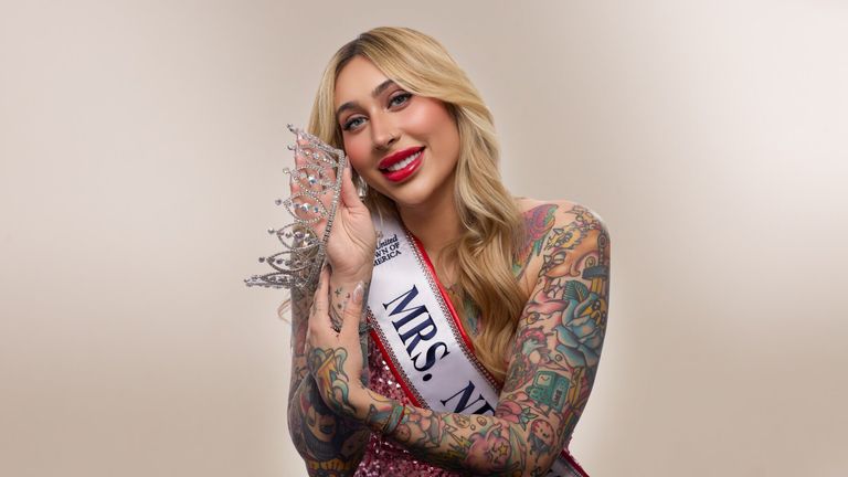 A smiling woman with long blonde hair and vibrant tattoos wears a sparkling crown and "Mrs. NY" sash, exuding confidence and joy against a neutral background.