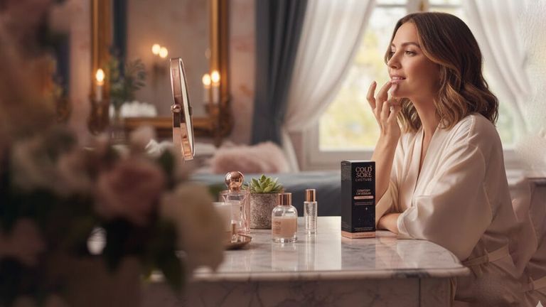 A woman applies lip care from Cold Sore Couture while sitting at a vanity, surrounded by elegant, luxury beauty products.
