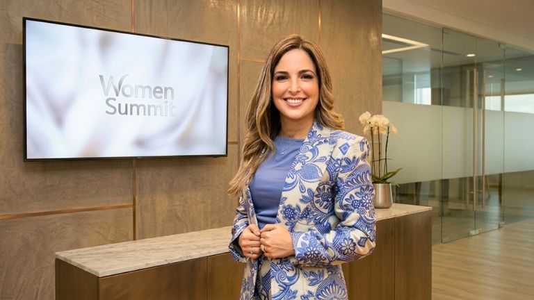 Ariana Acta stands before a large TV screen, representing her impactful journey from the Dominican Republic to global leadership.