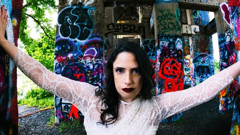 Woman in a white lace dress posing between colorful graffiti-covered concrete pillars.