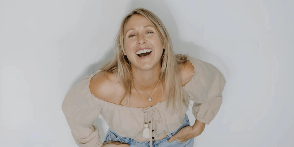 Libby Finlayson smiling with a joyful expression, wearing a beige off-shoulder blouse, and light blue jeans.