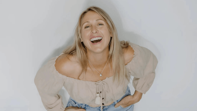 Libby Finlayson smiling with a joyful expression, wearing a beige off-shoulder blouse, and light blue jeans.