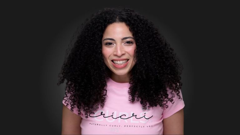 Smiling woman with voluminous curly hair wearing a pink Cricri Curls shirt against a dark background.