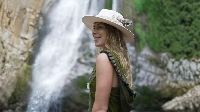 A woman stands by a waterfall, wearing a bohemian dress, wide-brimmed hat, and a serene smile, reflecting spiritual harmony.