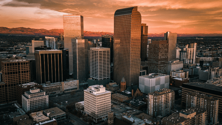 Aerial view of Denver skyline at sunset with downtown skyscrapers and Rocky Mountains in the background.