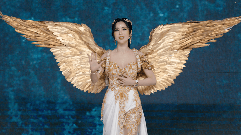 "Close-up of a model dressed in a glamorous angel-inspired costume with golden wings, giving a serene pose at a fashion show on a blue-lit stage."