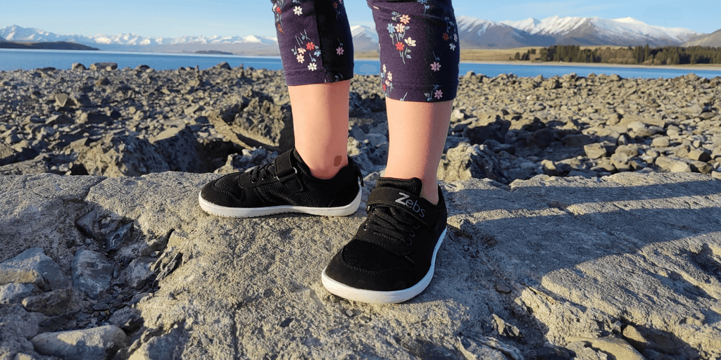 A child wearing Zebs Shoes, exploring rocky terrain by a lake with snow-capped mountains in the background.