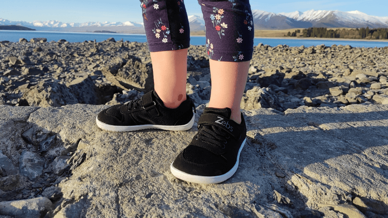 A child wearing Zebs Shoes, exploring rocky terrain by a lake with snow-capped mountains in the background.
