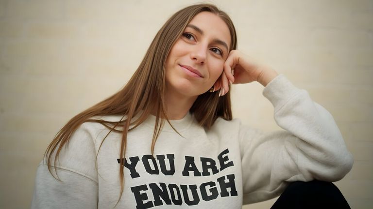 Young woman with long hair, wearing a "You Are Enough" sweatshirt, sits pensively with a gentle smile. The background is a soft, neutral color.