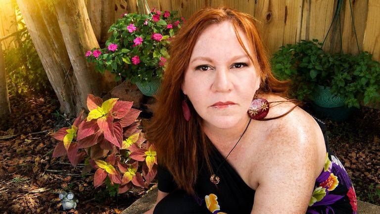 Red-haired woman seated in a sunlit garden surrounded by colorful plants and flowers, looking at the camera.