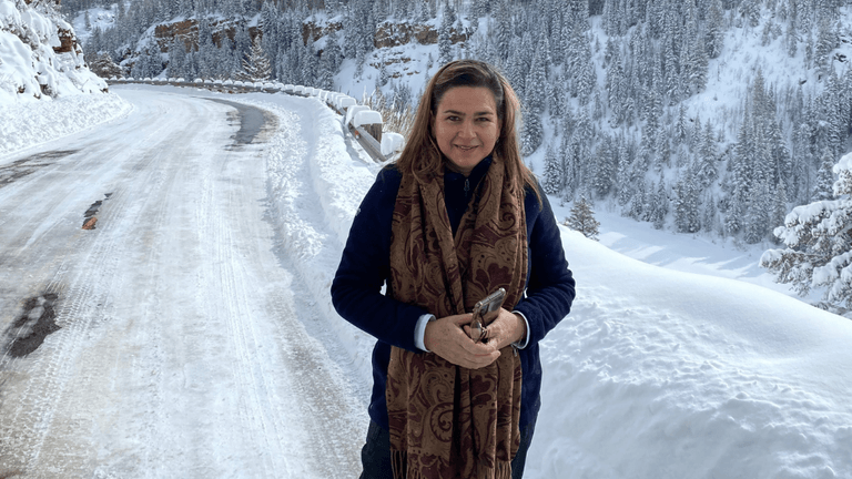 Woman holding phone on snowy mountain road with forested hills and winter landscape behind her.