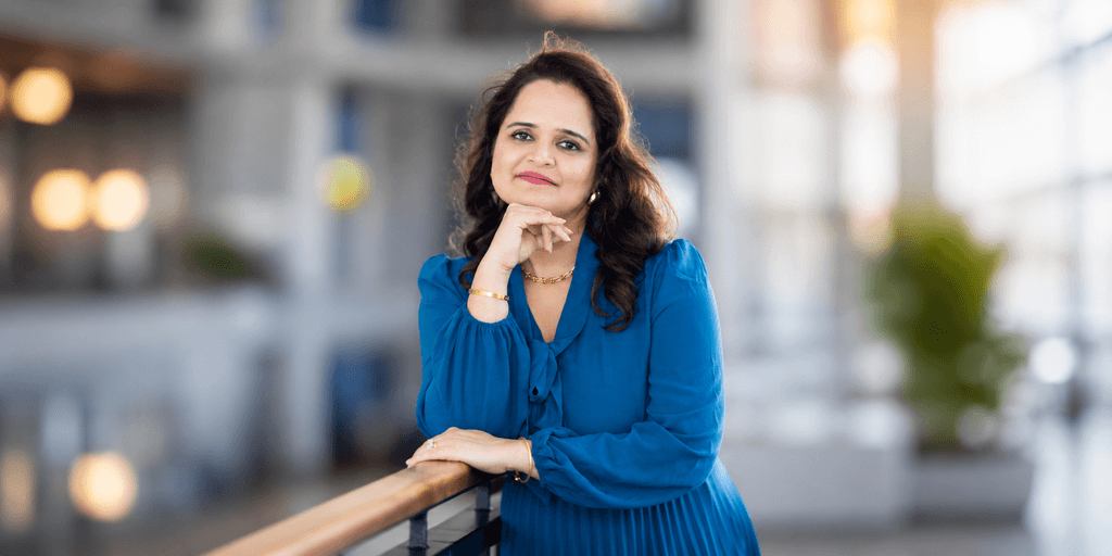 Isha Julka leaning on a railing, wearing a blue dress in a bright professional setting.