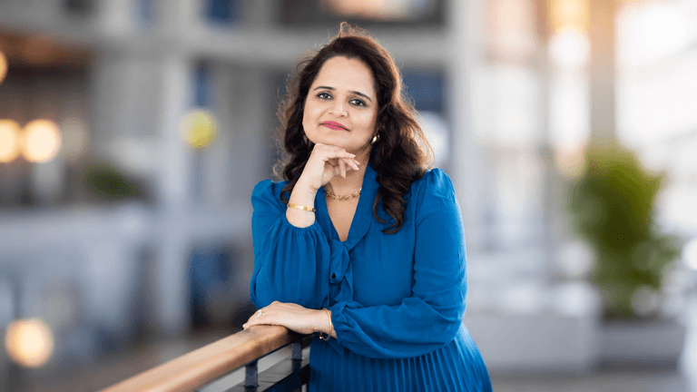 Isha Julka leaning on a railing, wearing a blue dress in a bright professional setting.