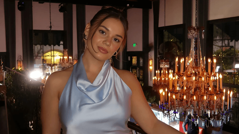 A woman in a satin blue dress smiles softly in front of chandeliers, exuding warmth and authenticity.