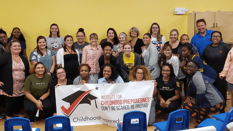 "A group photo of diverse individuals smiling and posing together with a banner from the Institute for Childhood Preparedness, promoting the message 'Don't Be Scared. Be Prepared'."