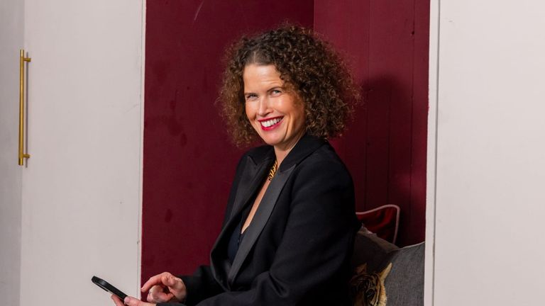 Dr Kate Armstrong smiling while seated with a smartphone, wearing a black blazer in a modern interior.