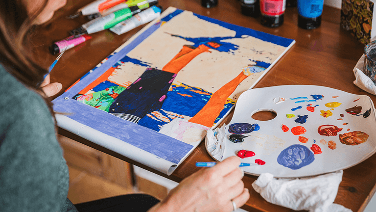 Artist painting a vibrant, colorful artwork at a desk with paint palette and supplies, reflecting creative expression.