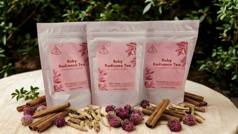 Three resealable pouches of Ruby Radiance Tea arranged on a wooden surface, surrounded by dried cinnamon, licorice root, and clover.