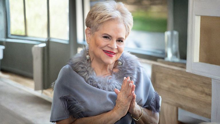 Elaine Adita Agnes smiling warmly, dressed in a chic, fur-trimmed gray shawl, radiating confidence and authenticity.