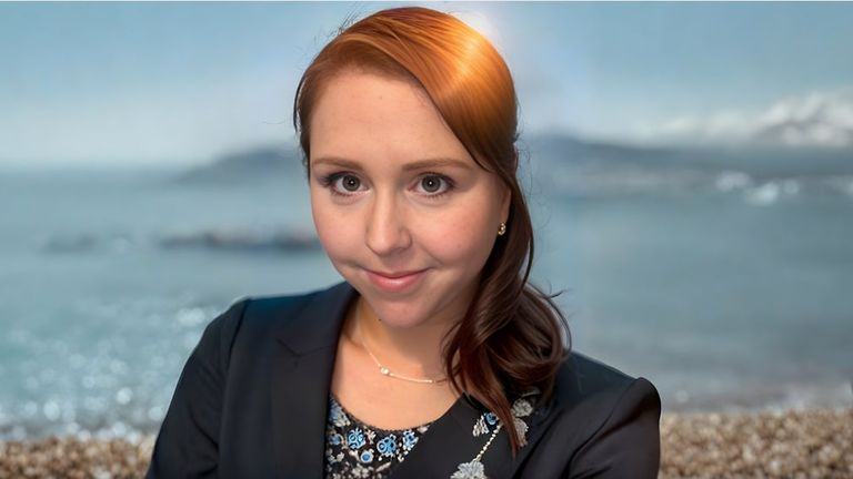 Professional portrait of Mel Schenker with auburn hair, a black blazer, and a serene ocean backdrop, exuding confidence.